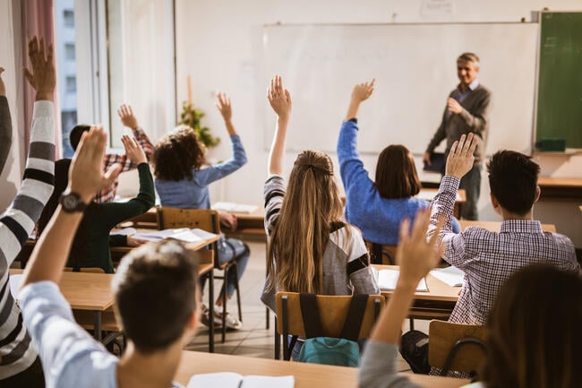 A classroom teacher stands at the head of a roomful of students who are raising their hands.