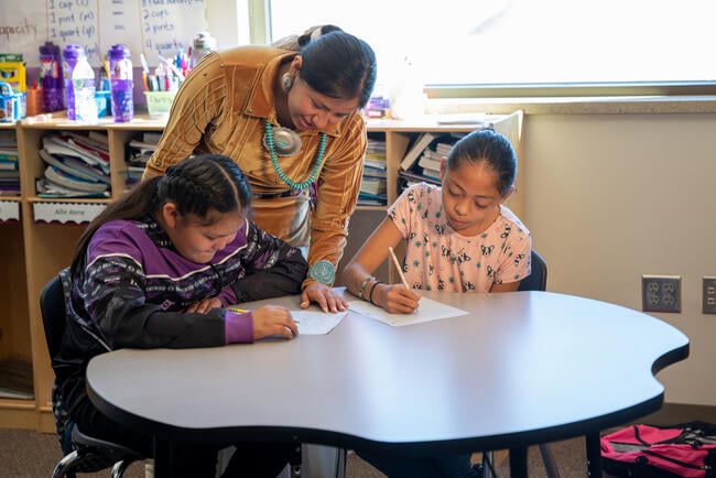 A teacher leans over a desk where two students are writing.