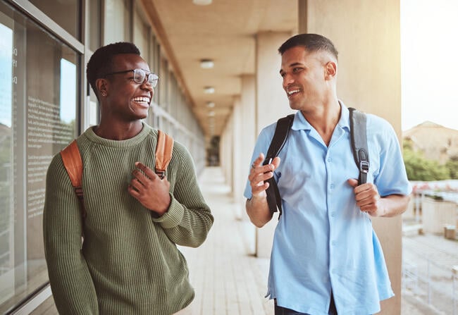 Two students wearing backpacks laugh while walking down a hallway at a college.