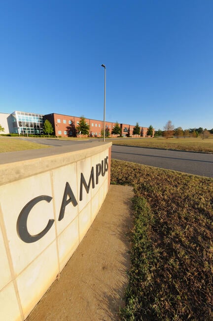 A wall with word “campus” written on it can be seen with an academic building and open space in the distance.