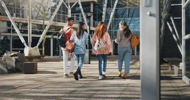 A group of four college students, each wearing backpacks, talk with one another while walking across campus.