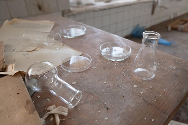 A photo of a dusty wooden table in an unused science laboratory; glass flasks, one knocked on its side, Petri dishes, and documents are strewn about the table.