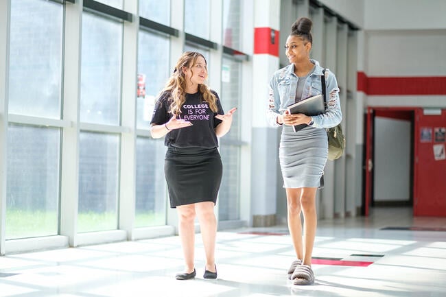 A shorter white woman wearing a shirt that says "college is for everyone" is shown talking with a taller Black girl in a denim jacket and skirt.