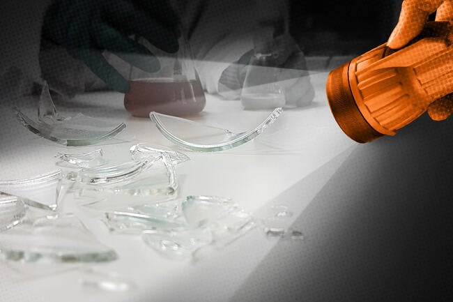 A flashlight shines a light on broken glass lab equipment.