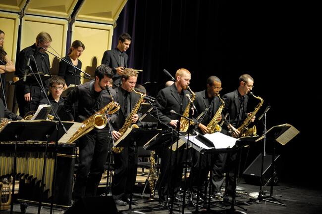 Five saxophone players stand to perform during a band concert. All students are clothed in black formal wear.