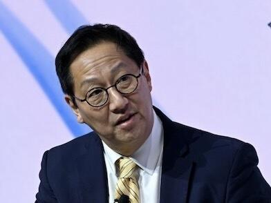 Santa Ono, an Asian man with black hair, wearing glasses and a dark suit with a yellow tie.