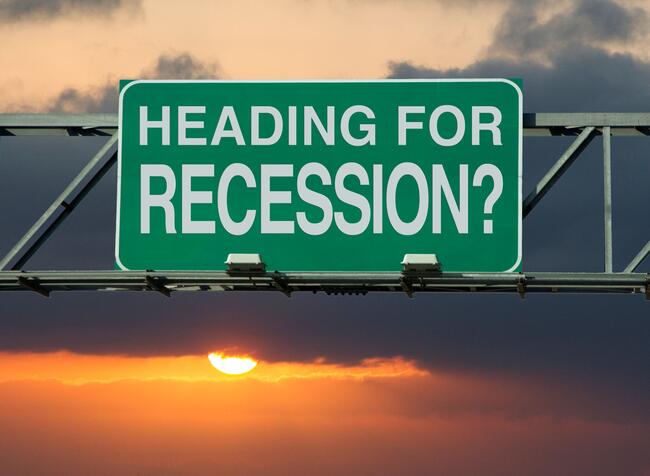 A green and white road sign, as if above a highway, says "Heading for Recession?" against the backdrop of a sunset in a cloudy sky.