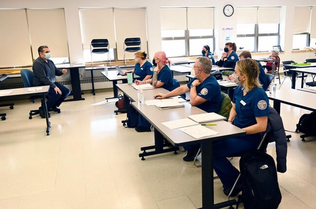 Students in scrubs and masks sit at desks listening to an instructor.