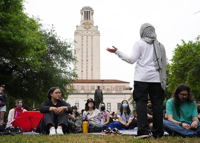 A woman wearing a kaffiyeh as a head scarf addresses a crowd of students seated in the grass in front of a building at the University of Texas at Austin.