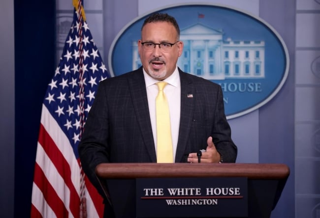 Secretary of Education Miguel Cardona, a middle-aged Hispanic man with a goatee wearing glasses and a business suit.
