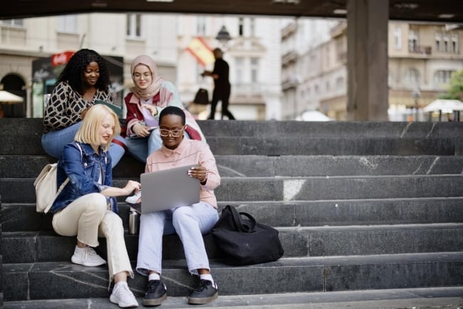 Students sit on the steps near the college and look at the laptop and digital tablet and talk