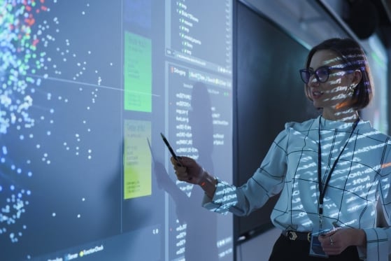 A woman points toward a screen filled with electronic blue dots. She also has electronic text projected onto her