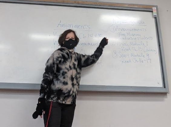 A professor teaching, wearing a mask, a parka and gloves.