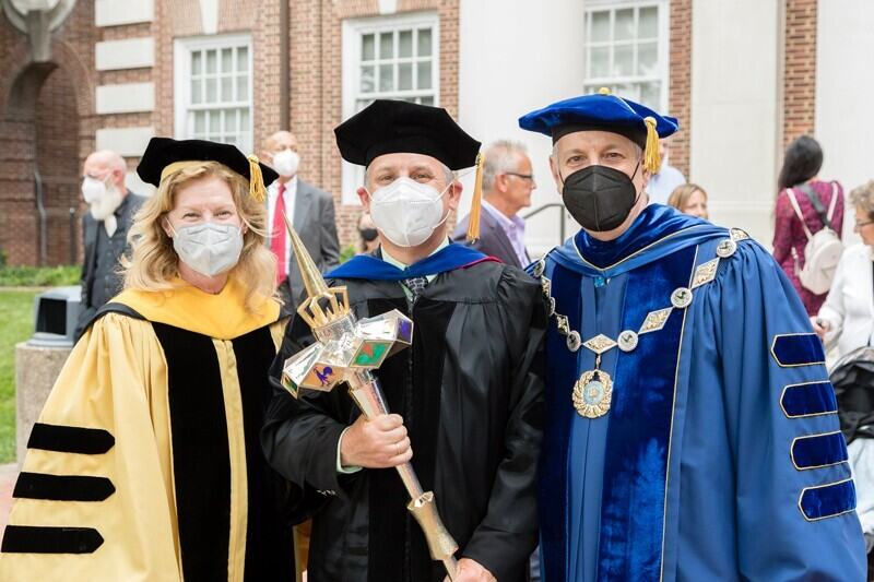 Provost Robin Morgan, Lou Rossi, dean of the Graduate College, and President Dennis Assanis, all wearing graduation robes and caps. Rossi is holding the university's mace.