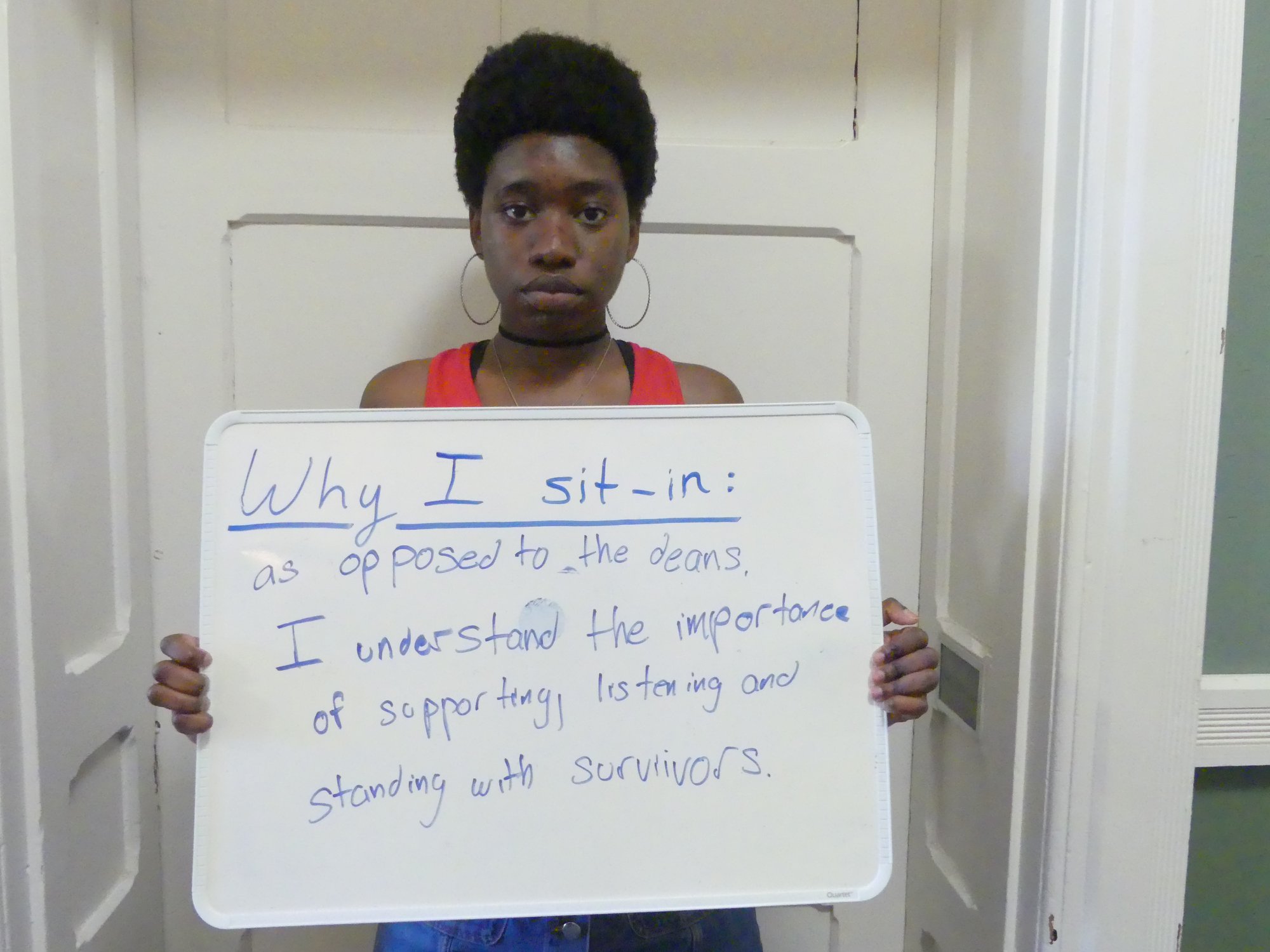 A black woman student holding a sign saying, "Why I sit-in: as opposed to the deans, I understand the importance of supporting, listening and standing with survivors."