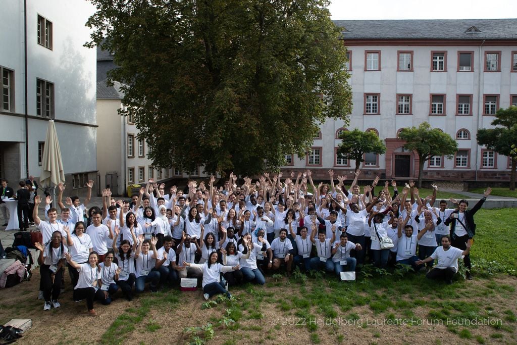 A large group of people, most of them wearing white shirts, on a patchy green lawn in front of white buildings.