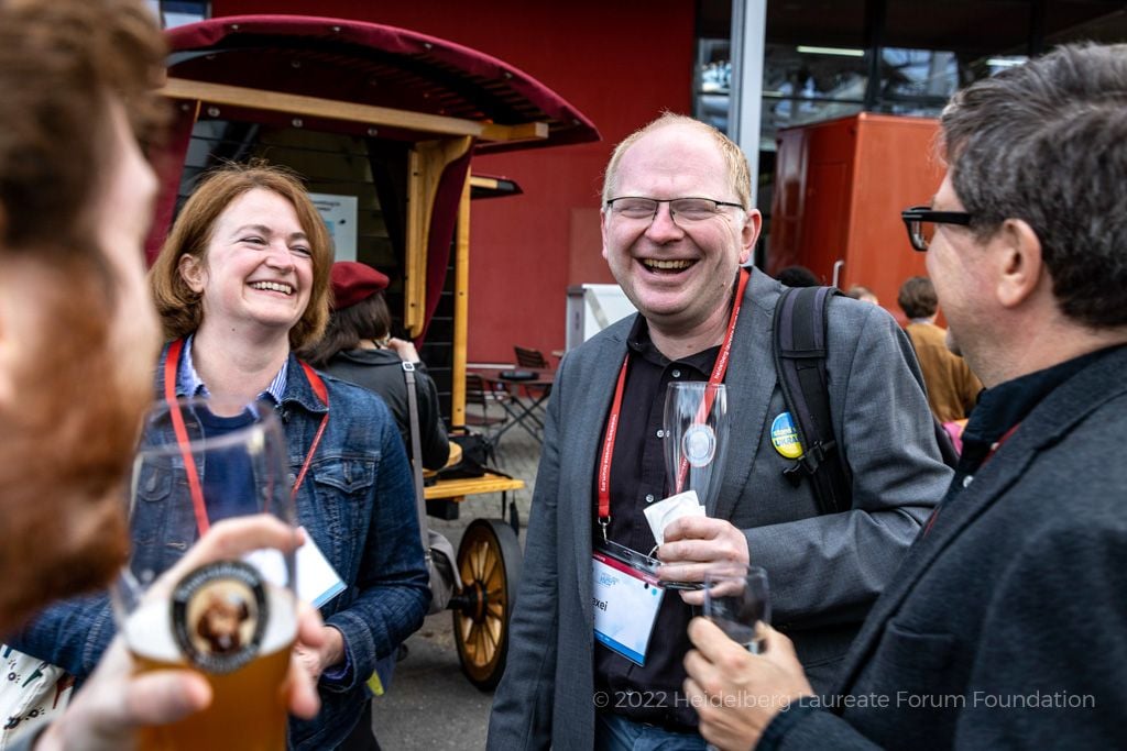 Four white people, one of them holding a tall glass of beer, and two of them wearing red lanyards signifying their participation in the summit, laugh together.