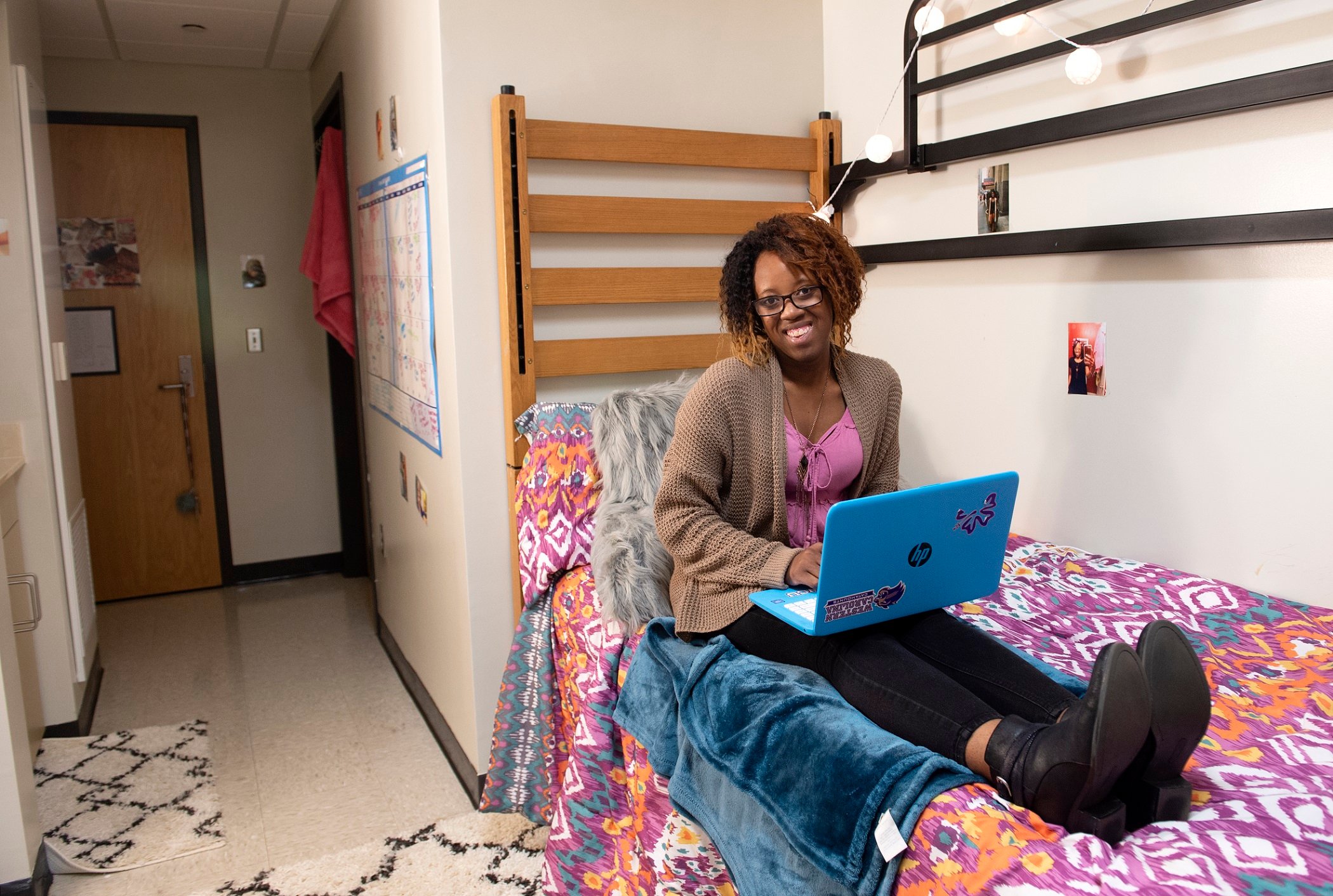 A young Black woman sits on a dorm room bed with a brighty colored duvet. She has a blue laptop computer on her lap.