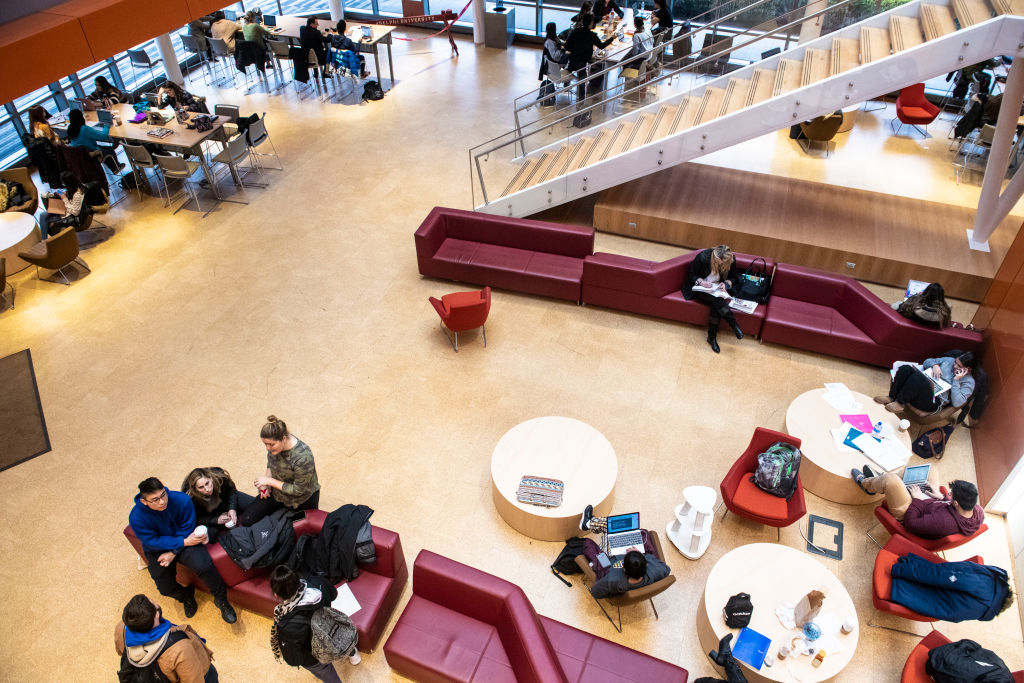 An open-plan gathering space in an academic building, with a floating staircase and tables and couches.