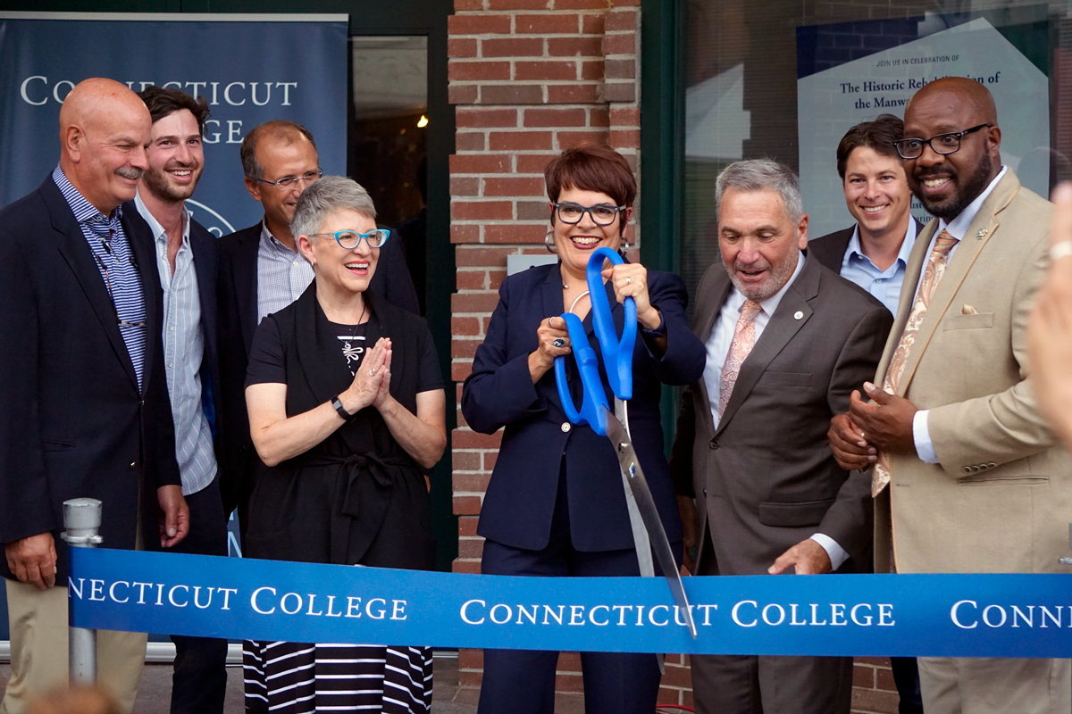 Katherine Bergeron, a light-skinned woman with short auburn hair and glasses, holds an enormous pair of blue-handled scissors for a ribbon cutting.