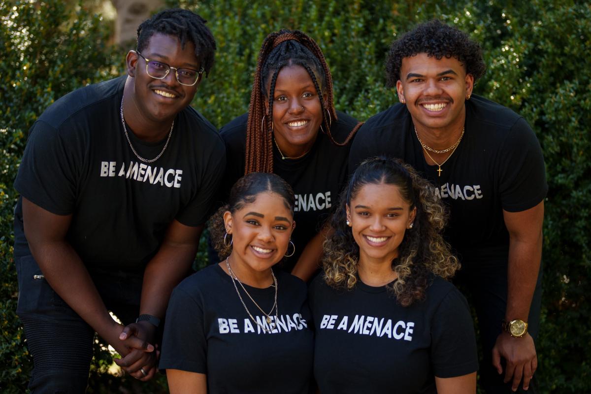 A group of students at Brigham Young University pose in matching black T-shirts that say "Be a Menace" in white, capital letters.