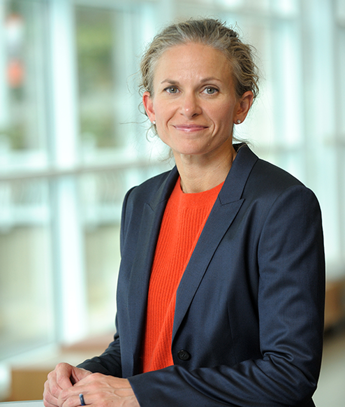 Brooke Elliott, a white woman with light hair wearing a blue blazer over an orange shirt.