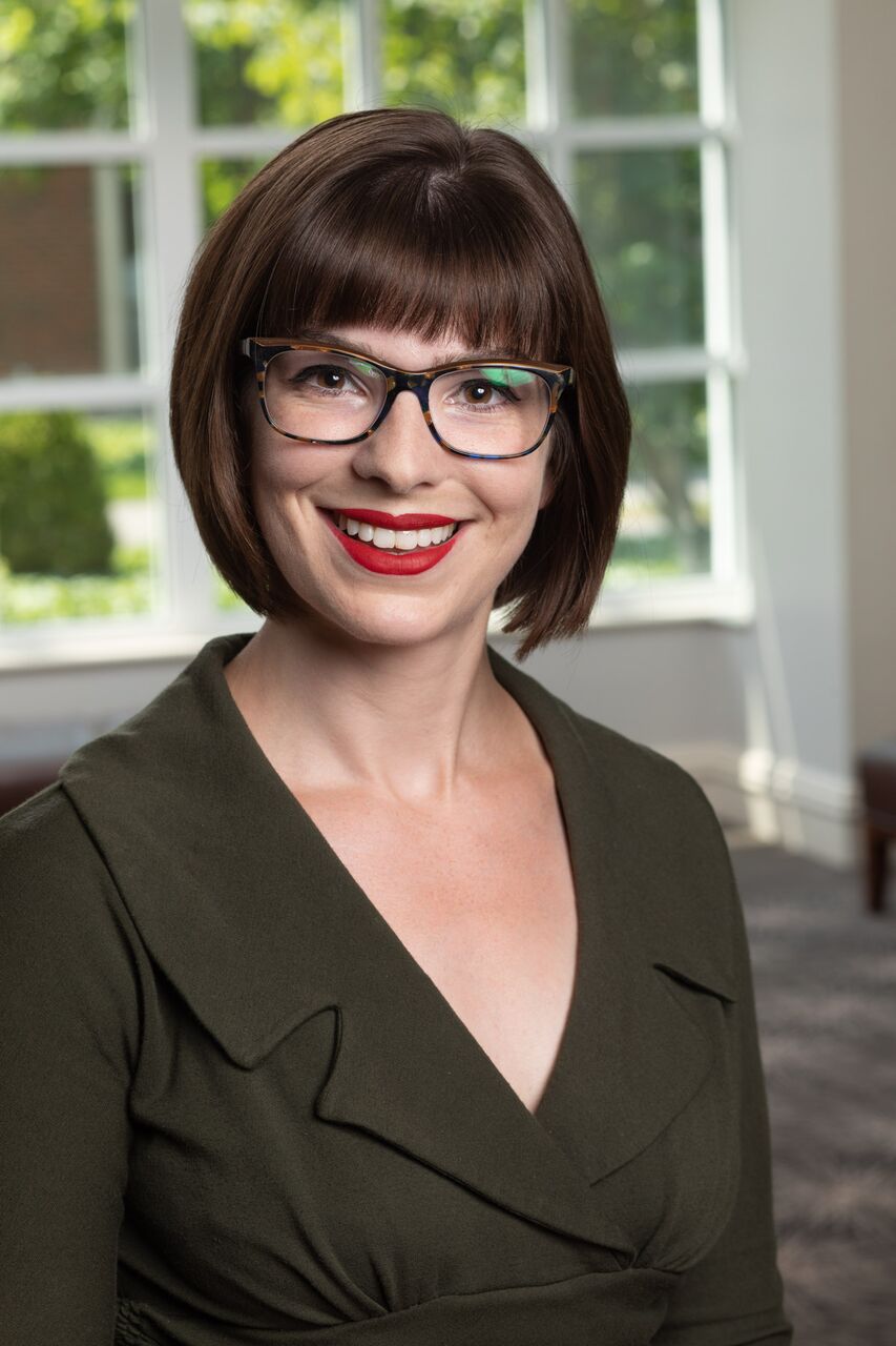 Kaitlin Dumont, a white woman with brown hair, glasses, and a very red lipstick.