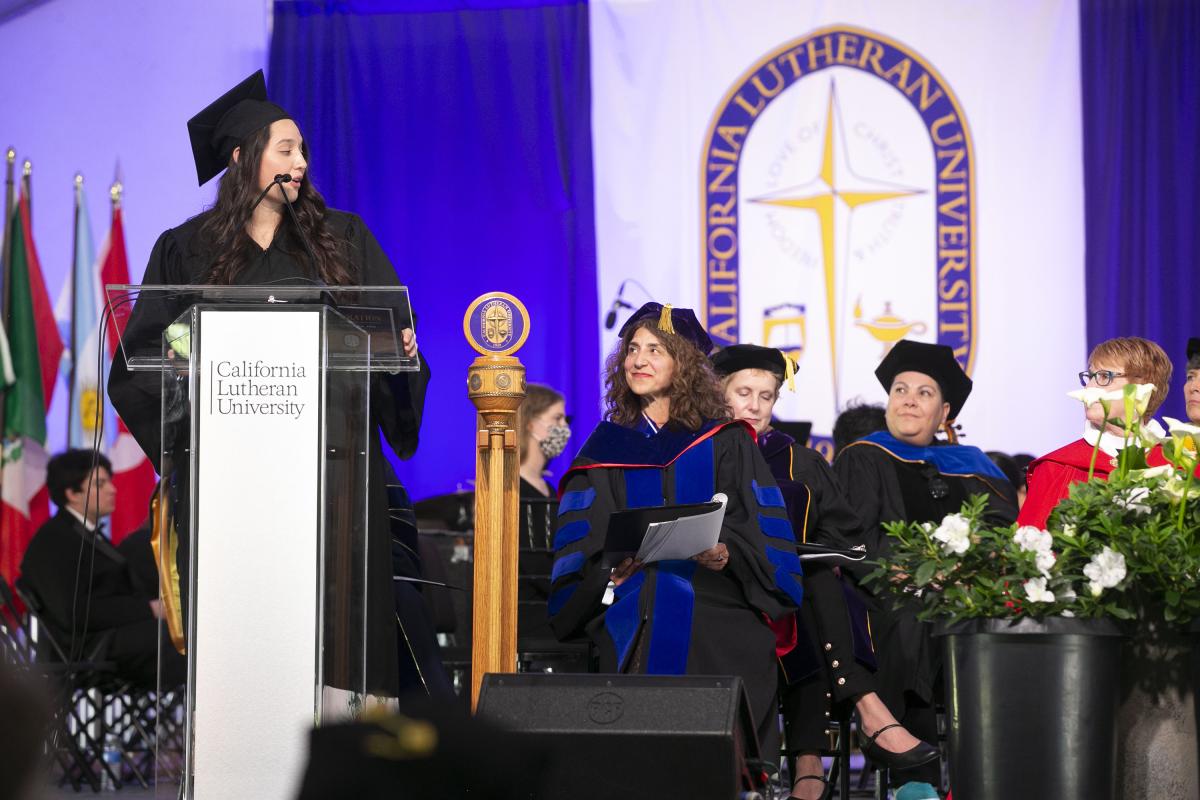 A dark-haired woman in academic cap and gown at a podium with "California Lutheran University" on the front. The president of the university, a woman with dark hair, is seated to her left.
