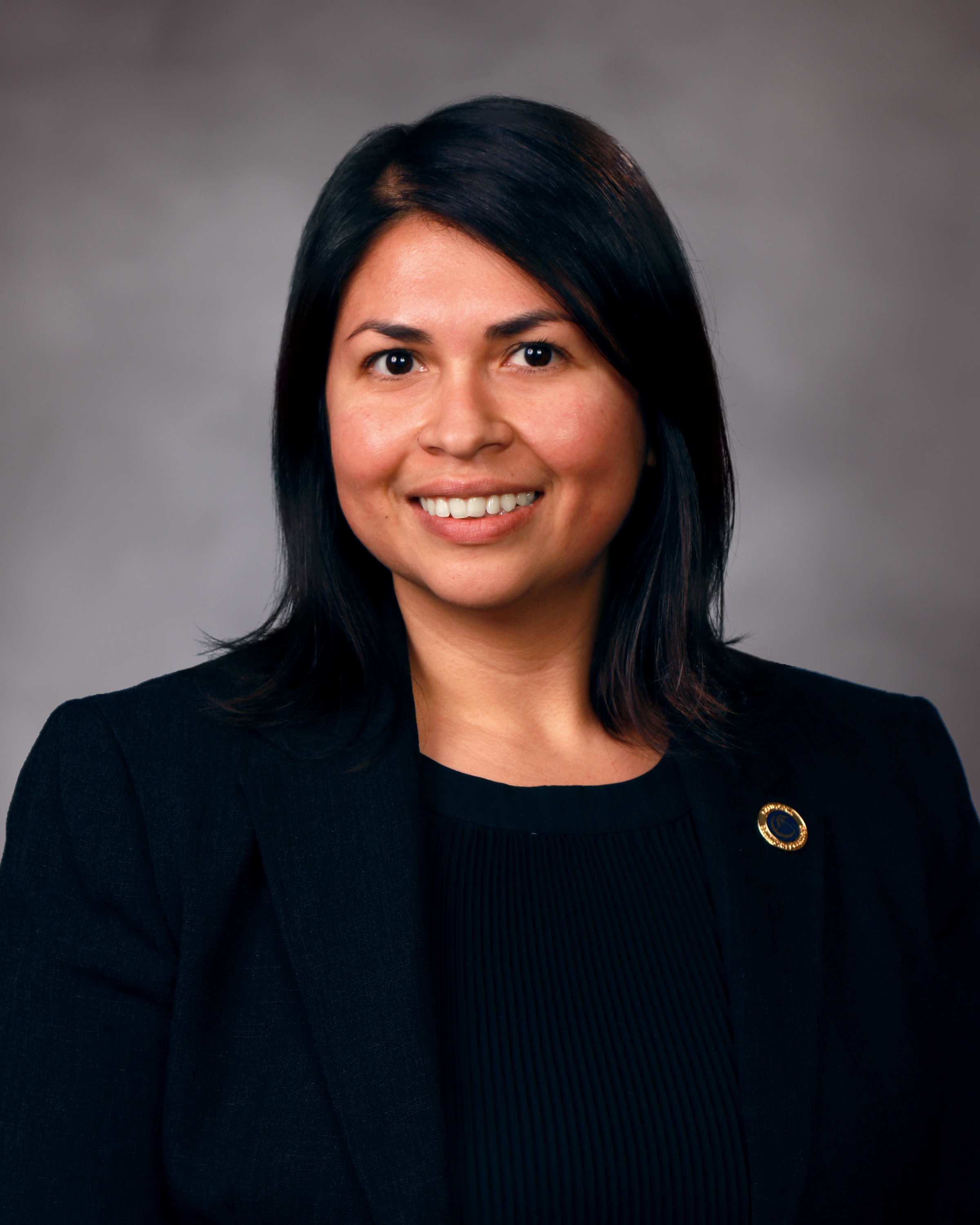 Daisy Gonzales, a Latina woman with dark hair who is wearing a black top with a gold pin.