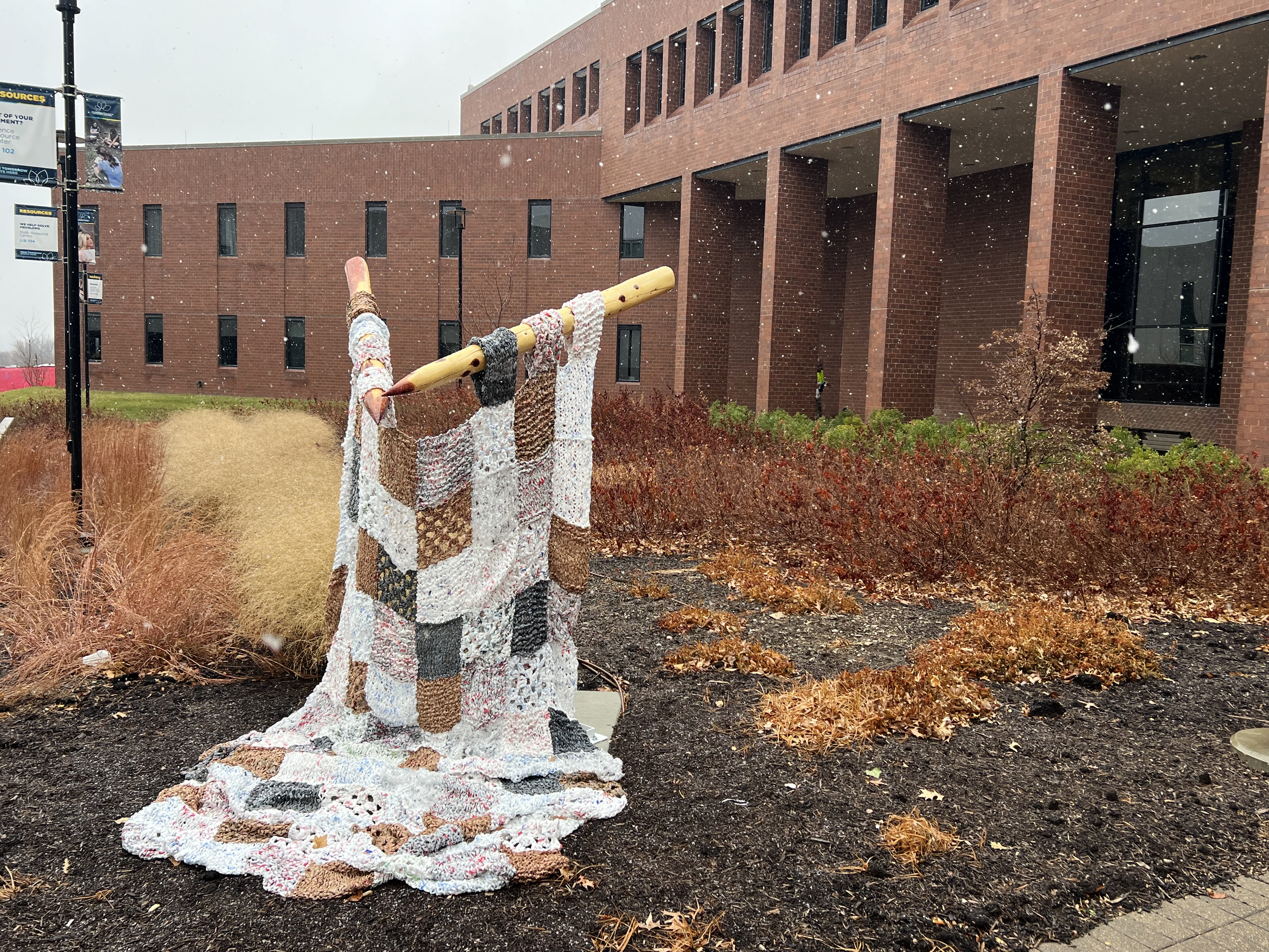 A sculpture made of recycled plastic bags outside an academic building. The sculpture looks a lot like a piece of knitting on large wooden knitting needles.