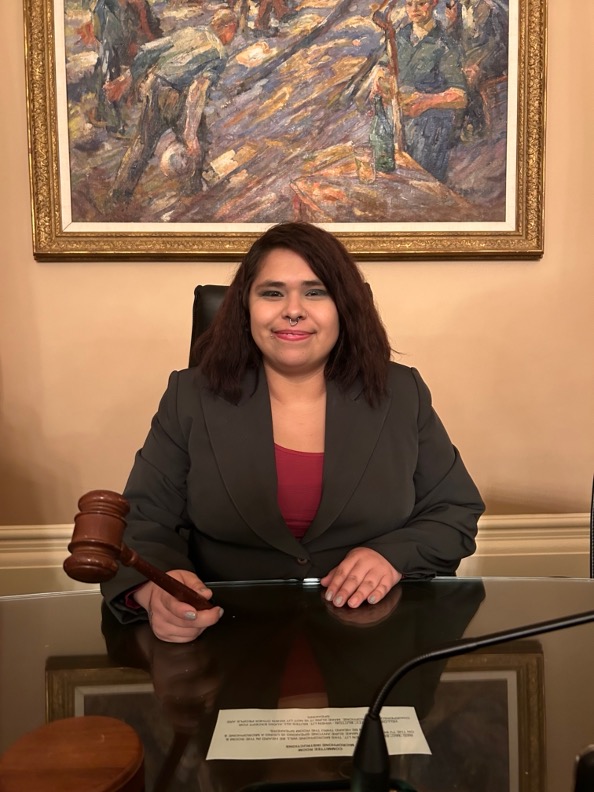 Christina Torres, a young woman with dark hair and light brown skin, wearing a nose ring and pink lipstick. She is holding a gavel and sitting at a microphone.