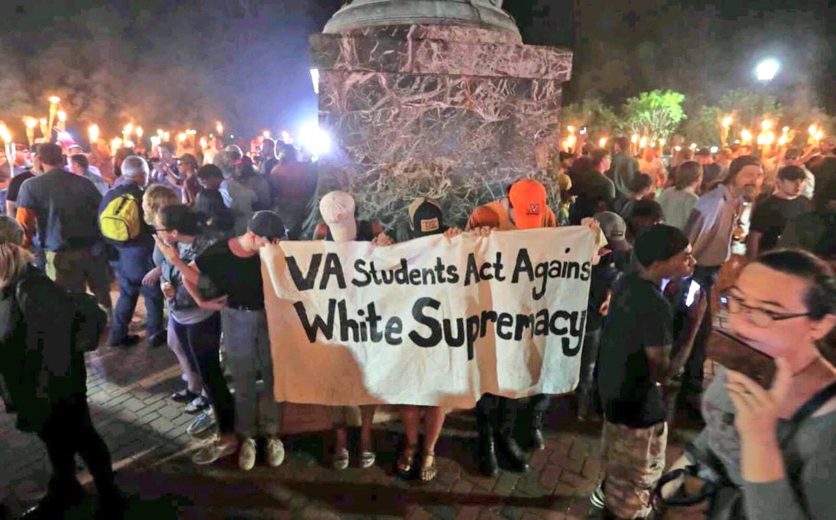 Counterprotesters against a white nationalist gathering hold a sign saying "VA Students Act Against White Supremacy." The counterprotesters are at the foot of a statue of Thomas Jefferson and are surrounded by torch-bearing white nationalists.