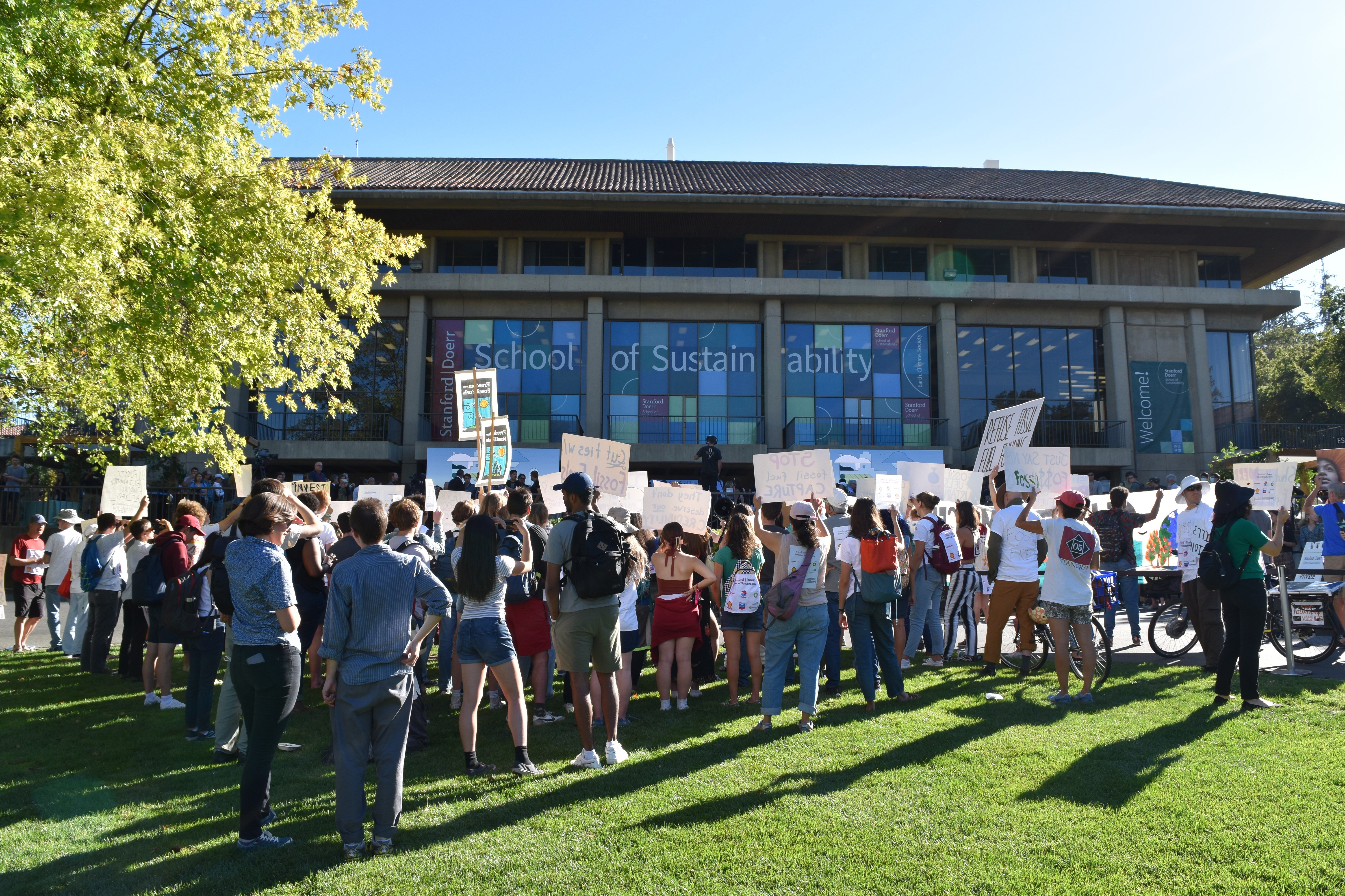 A group of people with signs and backpacks in front of a building that says "School of Sustainability."