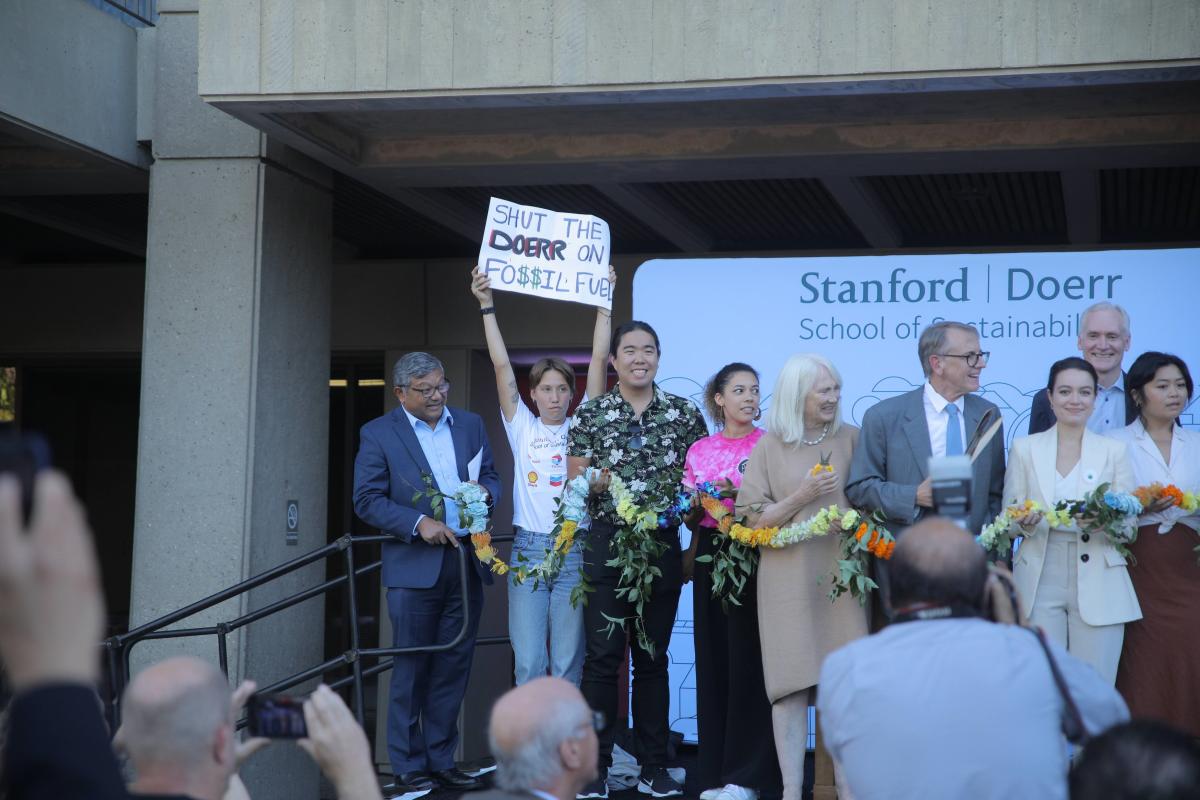 A protester hoists a sign on stage next to smiling people in suits and dresses. The sign reads "Close the Doerr on Fossil Fuels."