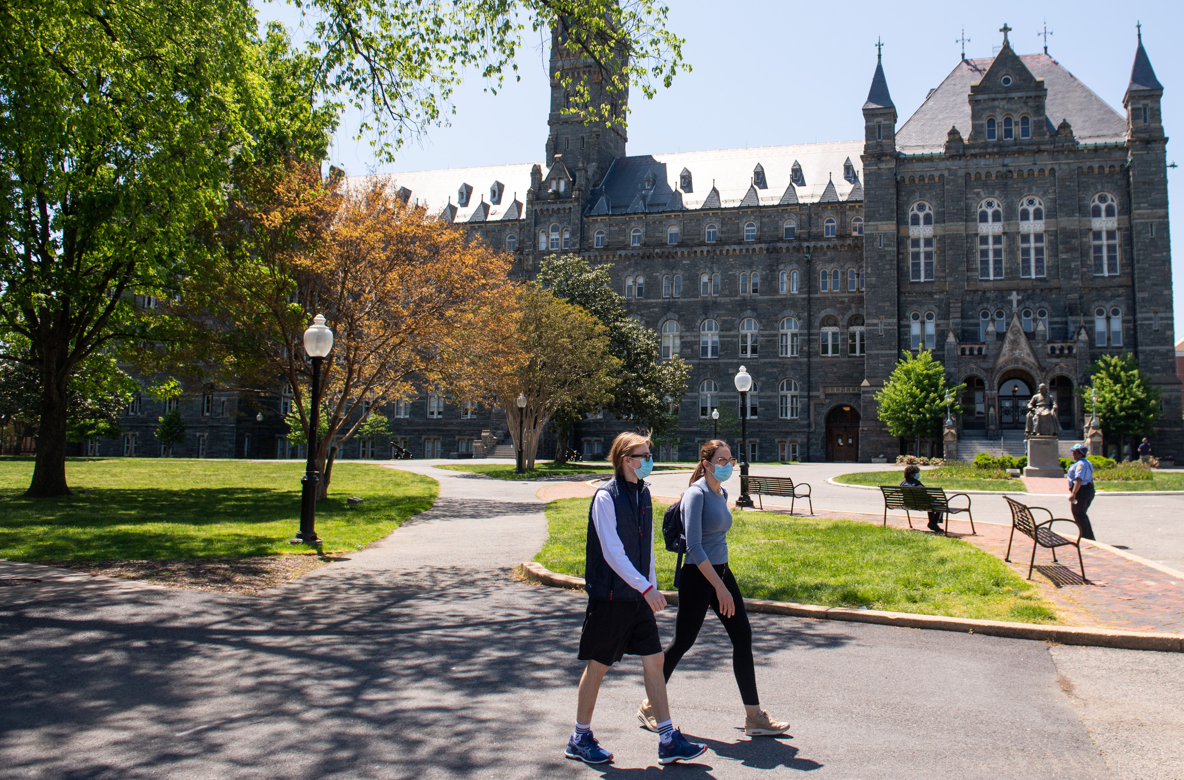 Maskeli öğrenciler süslü bir akademik binanın önünde yürüyor.