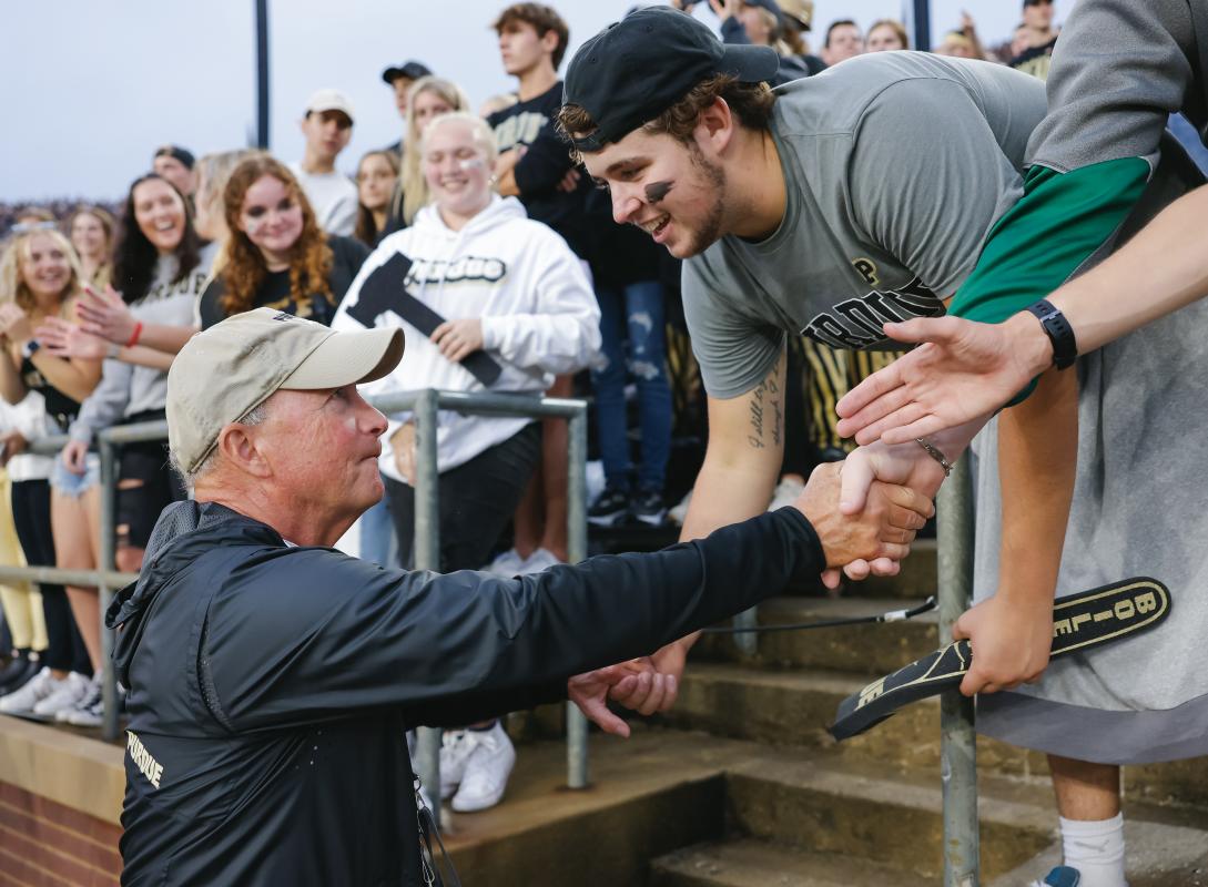 Mitch Daniels, an older white man in a baseball cap, greets Purdue students at a sporting event.