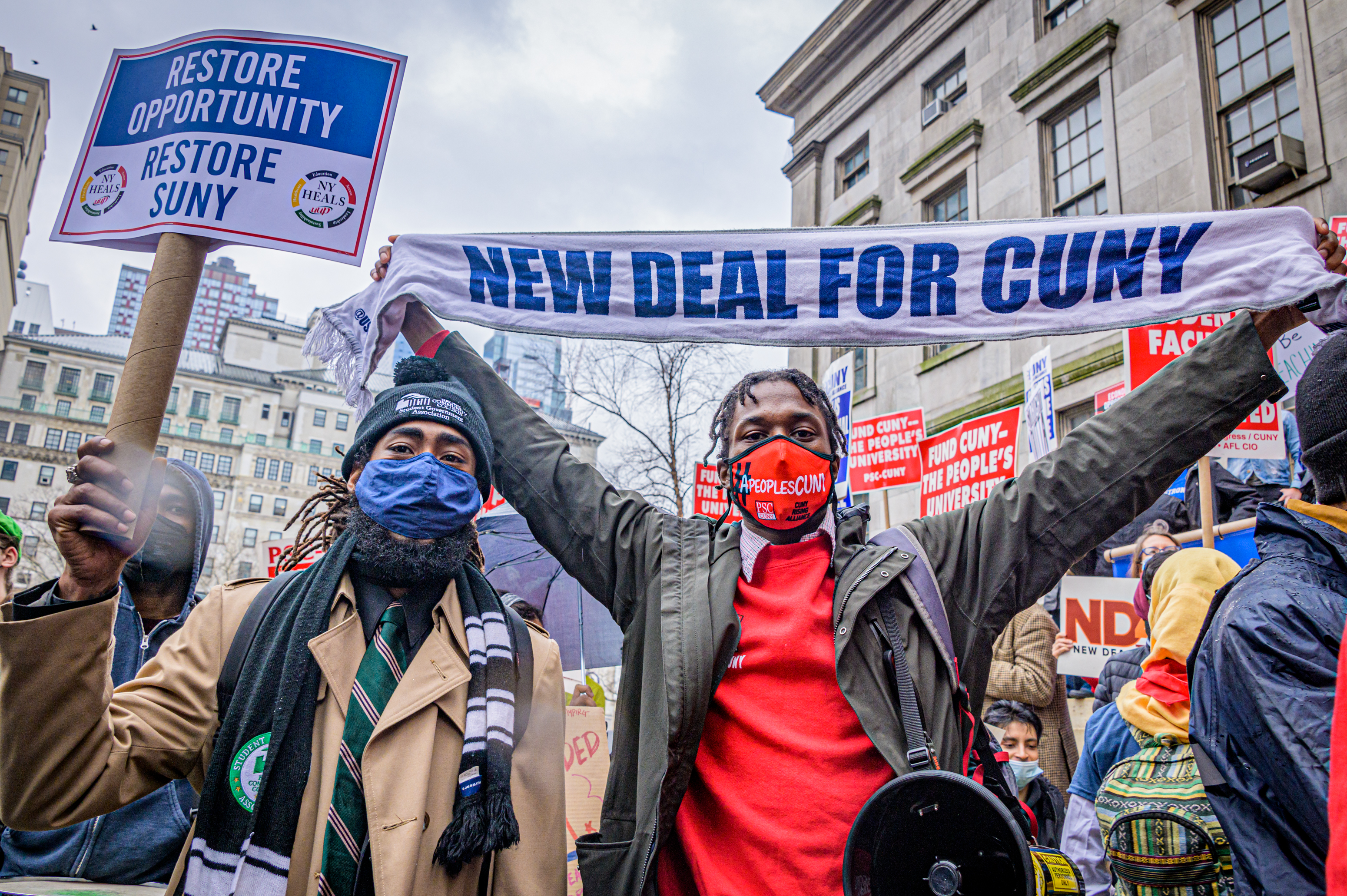 Two protesters, both Black men, hold signs demanding the New York State government boost funding for SUNY and CUNY, in the middle of a crowd of other protesters.