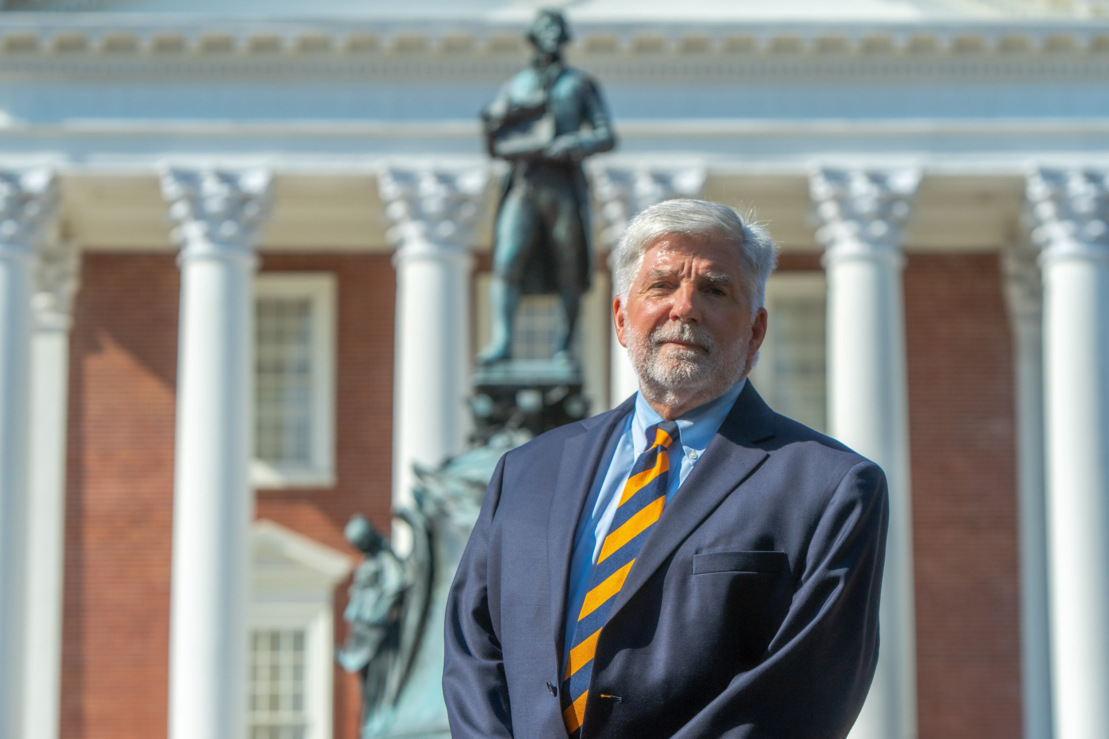 Bert Ellis, a white man wearing a suit and a tie in the blue and orange colors of the University of Virginia, stands in front of a statue of Thomas Jefferson.