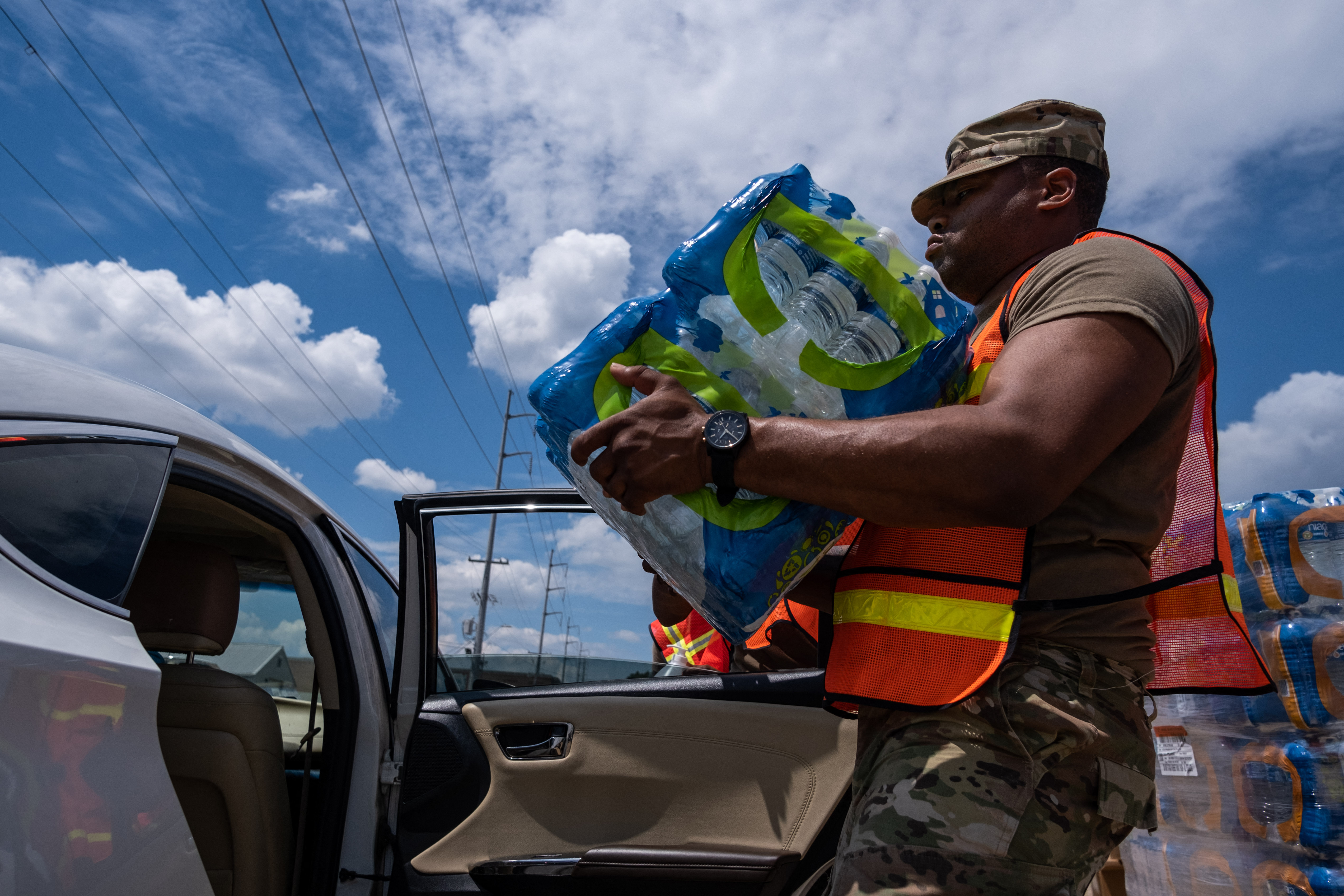 A Black National Guardsman wearing a high-visibility vest carries a case of bottled water.