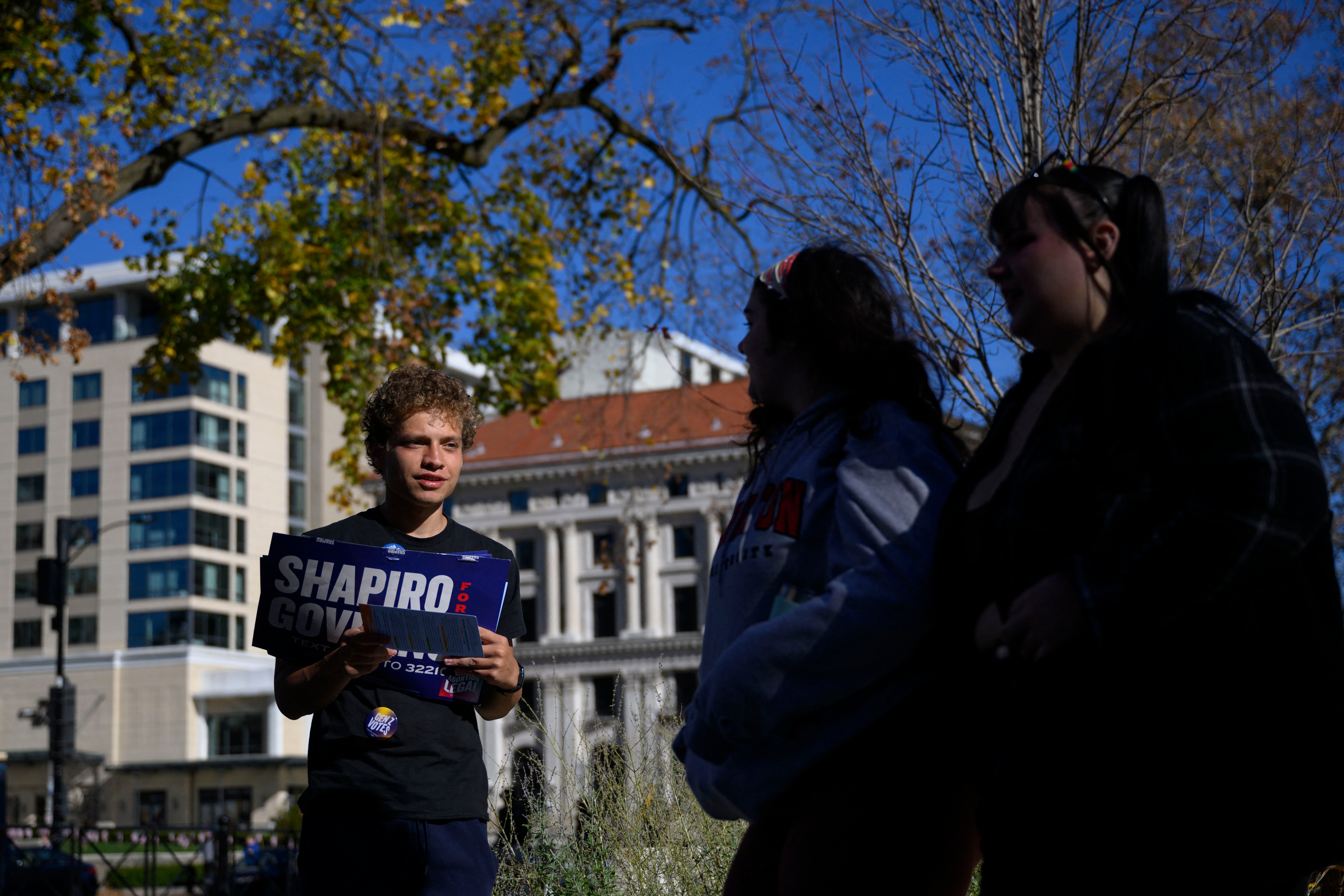 A young man holds a "Shapiro for Governor" sign as he speaks to two young women in front of a tree branch and buildings in Pittsburgh.