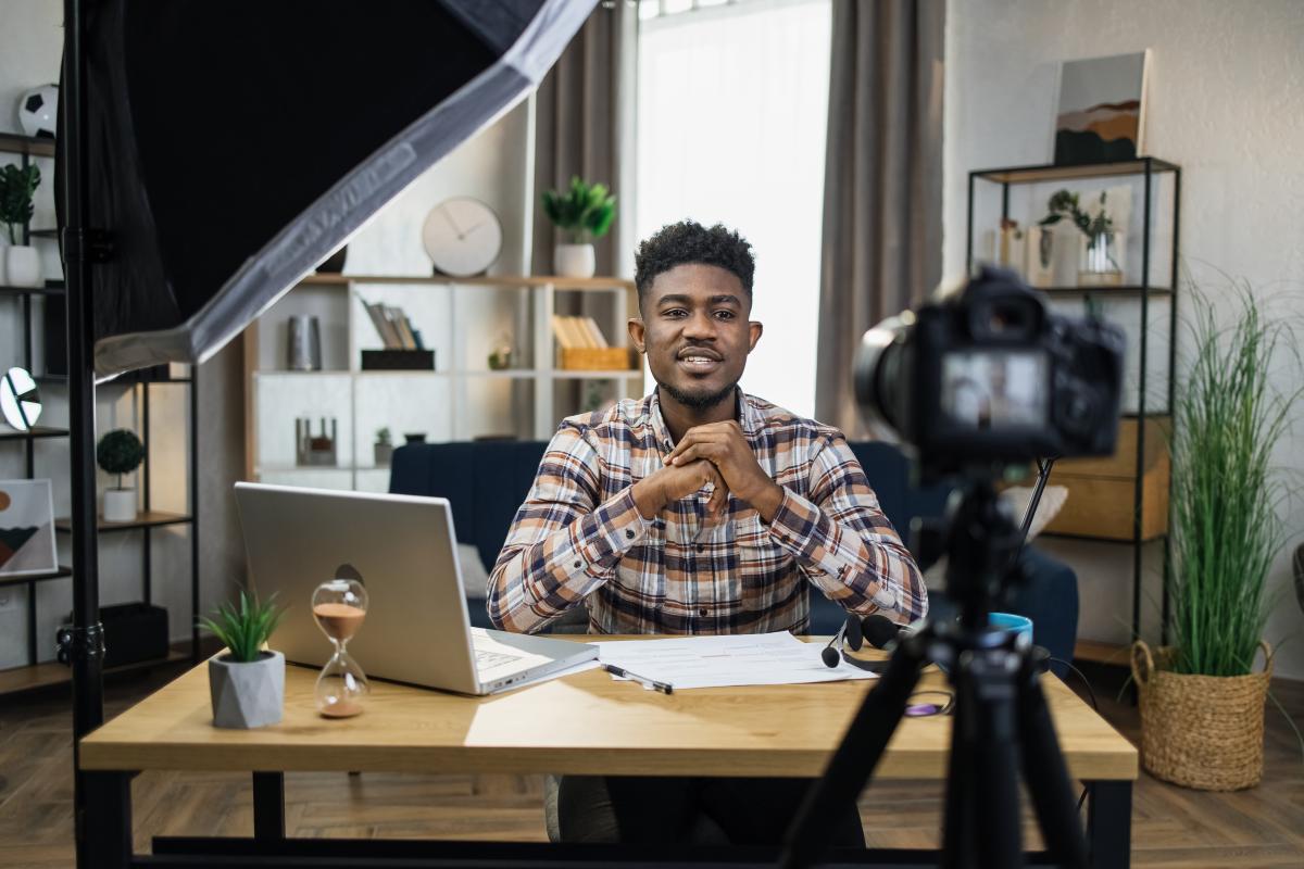 A young Black man sits at a desk with a laptop open next to him. There is a camera pointed at him.