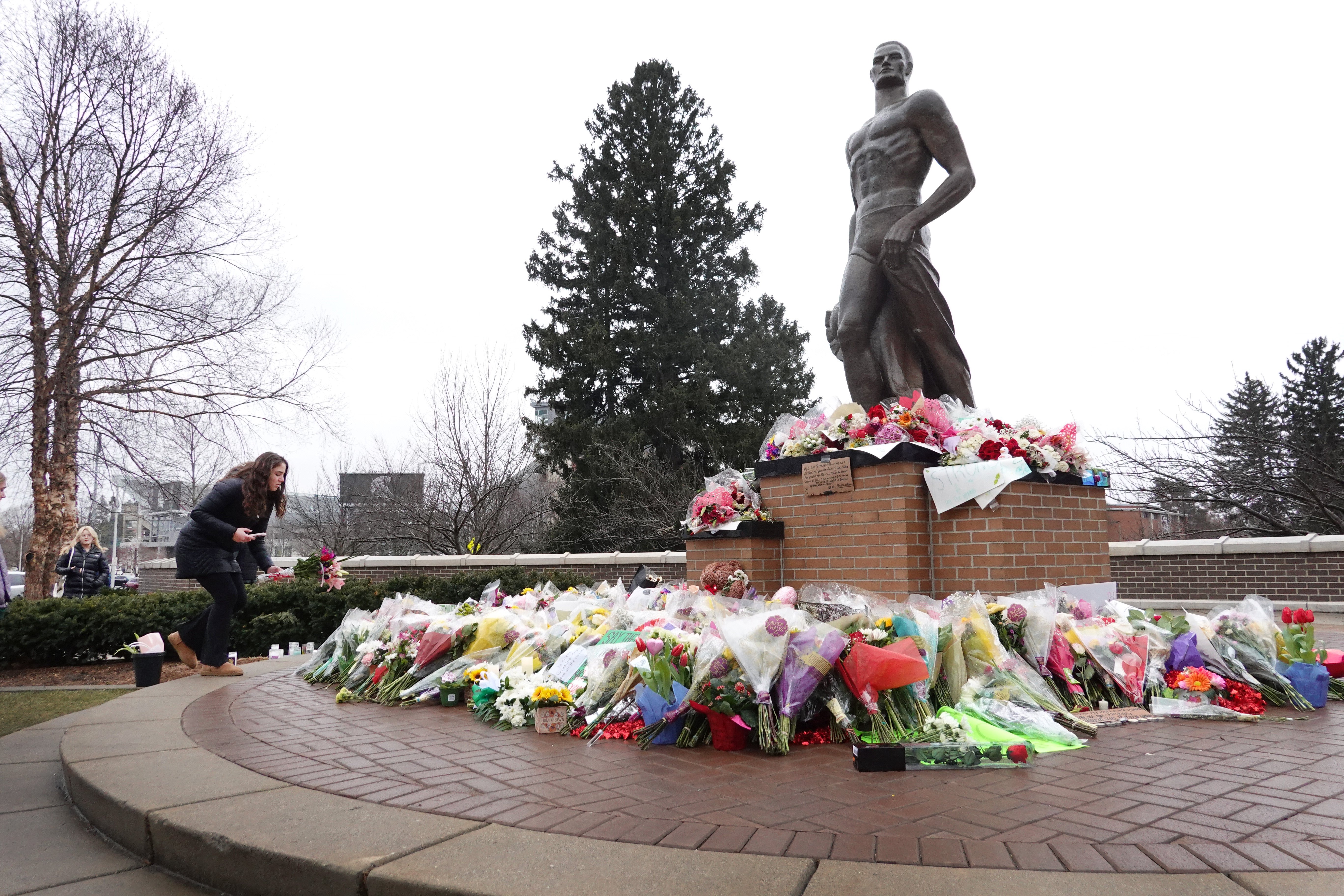 a woman lays a bouquet of flowers with a large assemblage of other bouquets at the feet of a muscular male statue on a brick podium