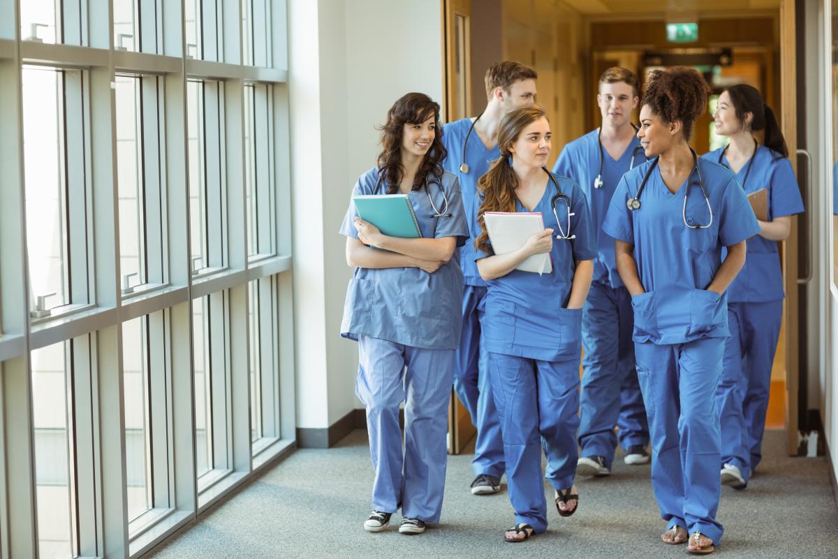 A diverse group of medical students in blue scrubs walks down a hallway.