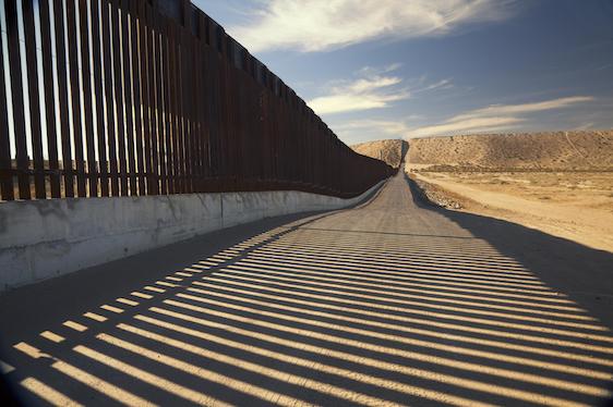 Border wall in the desert with shadows, blue sky and clouds.