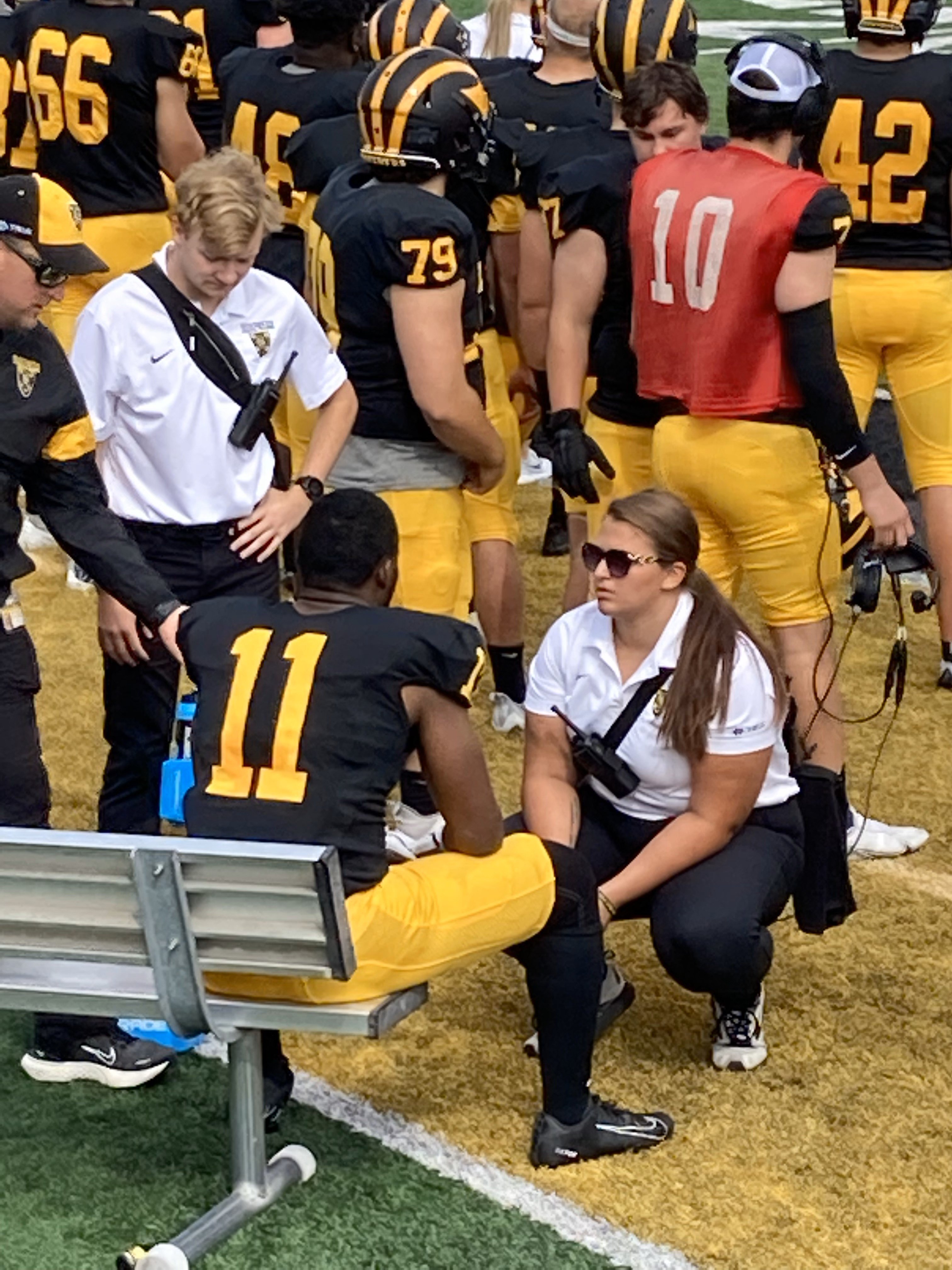 Afemale athletic trainer works on the sideline of a football field with football players in black and gold uniforms.