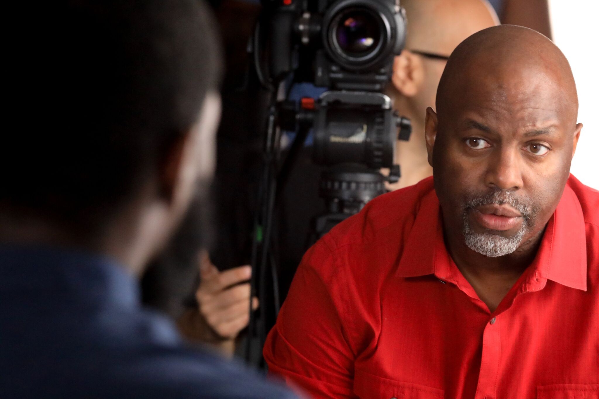 Byron Hurt, a Black man with a goatee wearing a red shirt, faces the camera, conducting an interview. The person he is interviewing has their back to the camera.