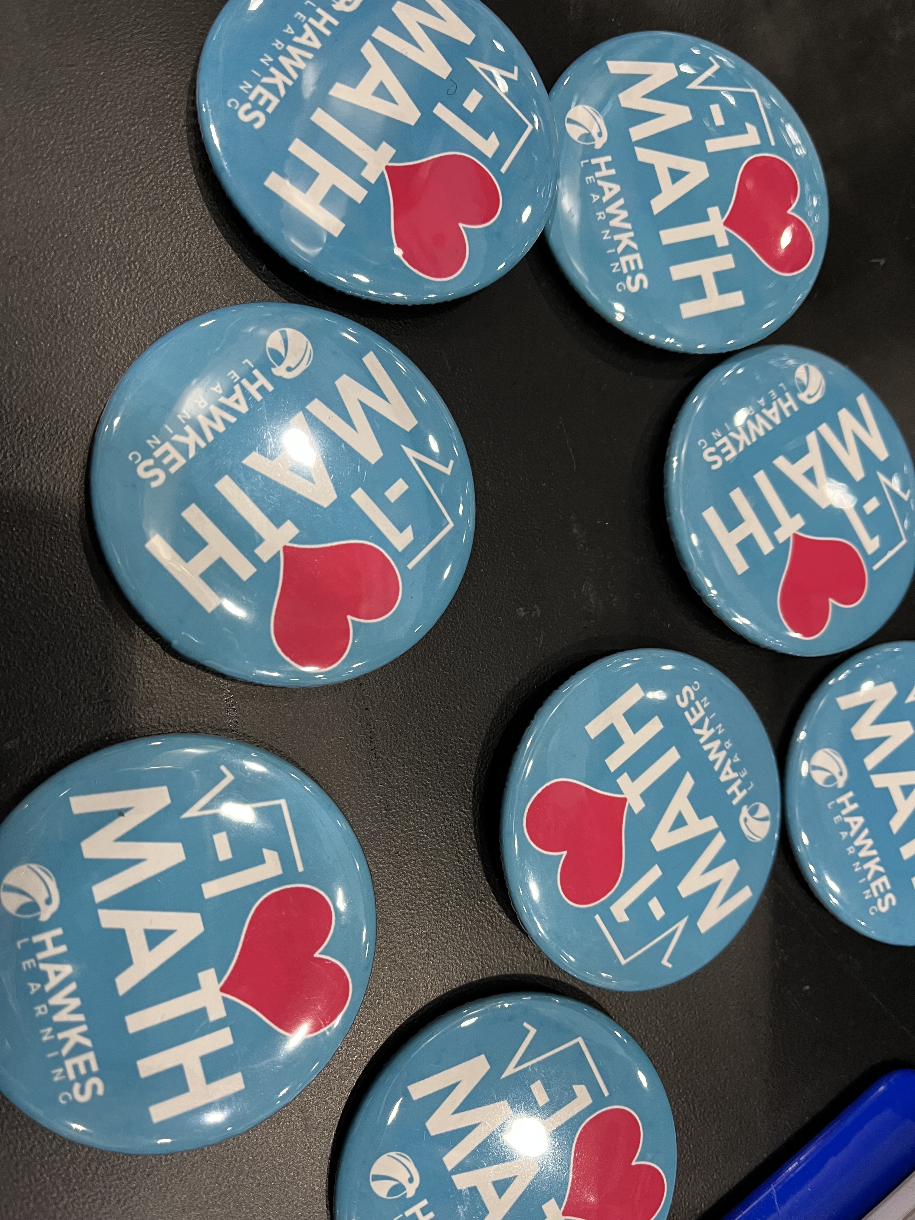 Numerous buttons on display on a table at the Joint Mathematics Meetings. The buttons are alike and use mathematical symbols to express “I love math.” The square root of negative one is used in place of “I.” A red heart is used in place of “I.” In small print at the bottom of each button are the words, “Hawkes Learning.” 