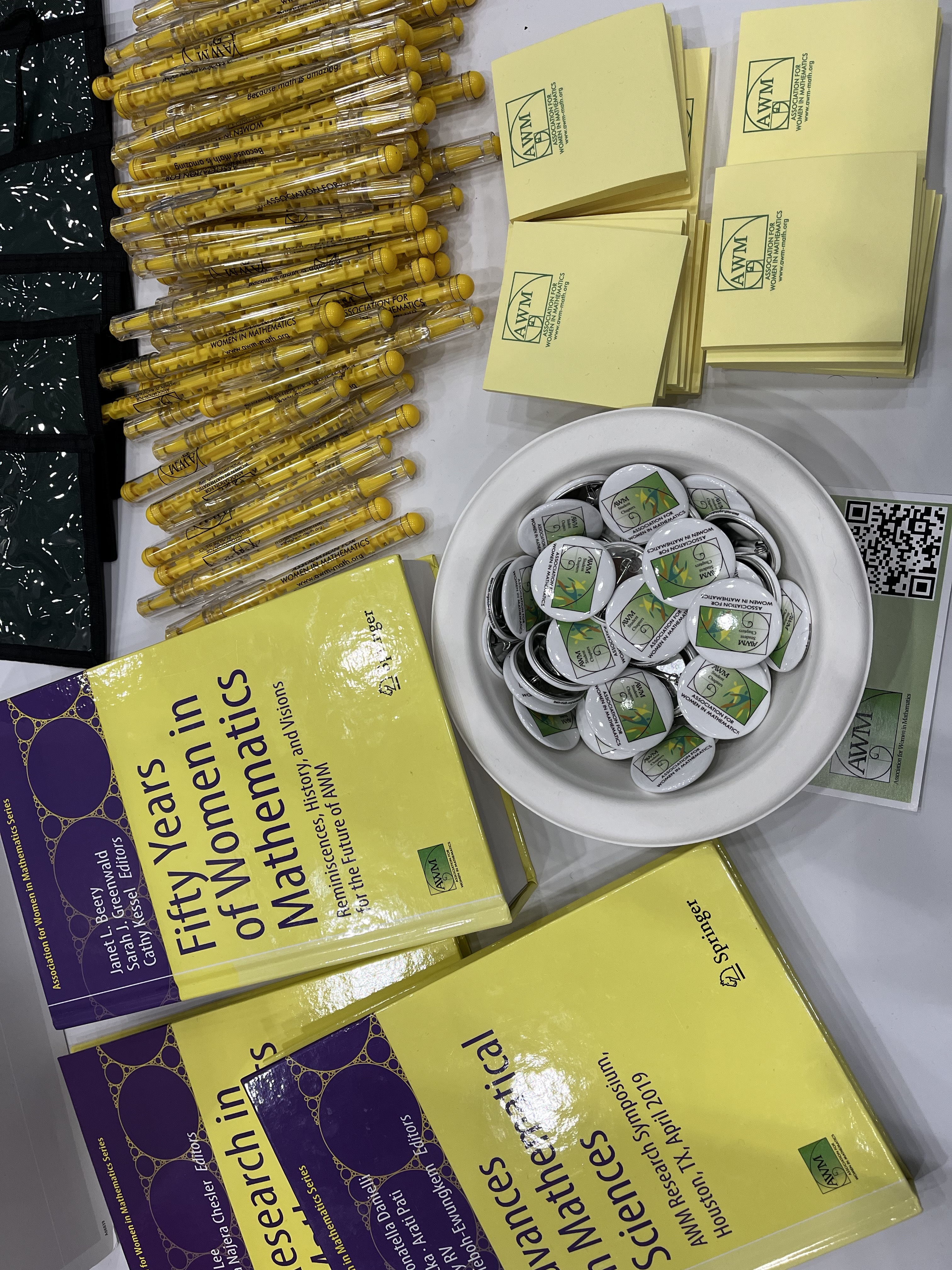 A table in the Joint Math Meetings exhibit hall with items from the Association for Women in Mathematics. The items include three books from the association, including one titled, “Fifty Years of Women in Mathematics.” There are also items with the association’s logo, including pencils, buttons, and Post-its. 