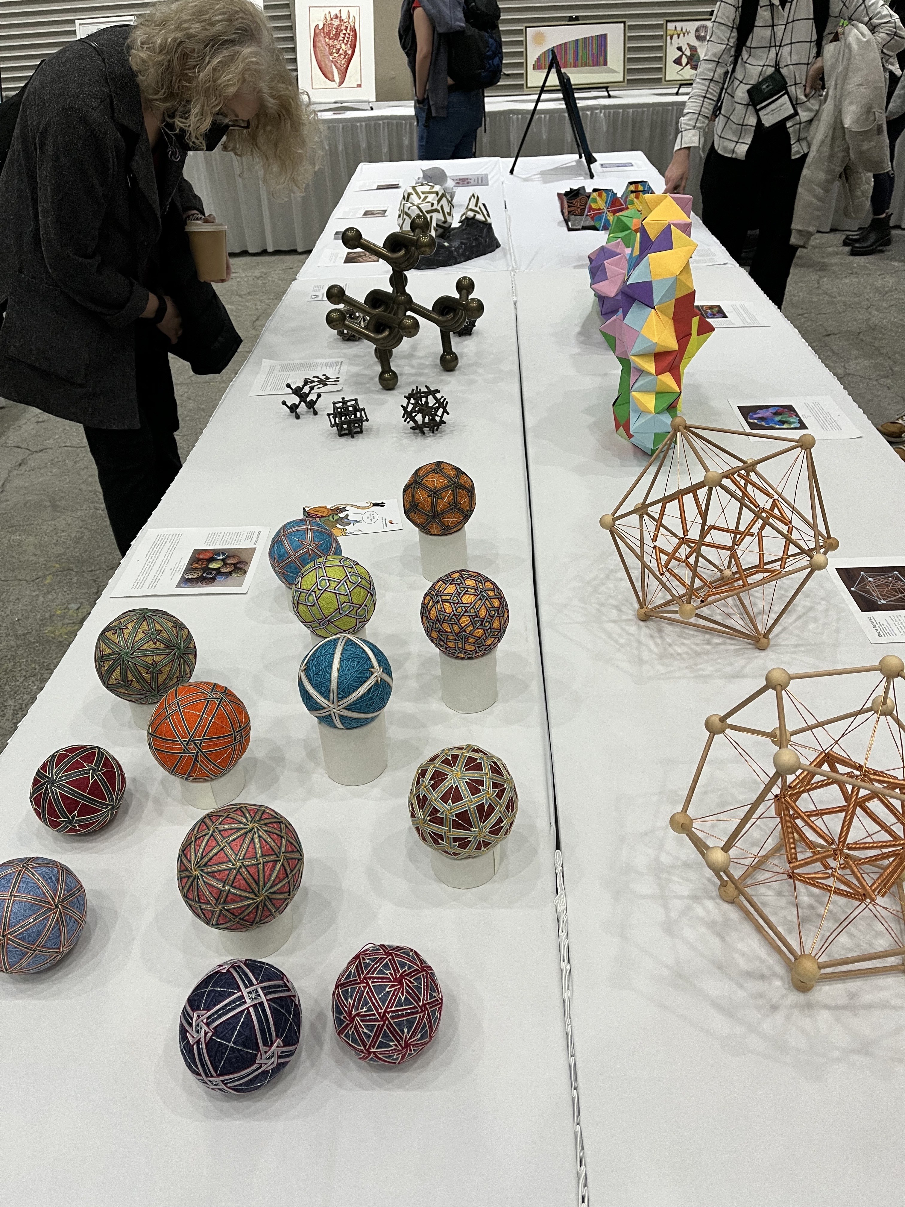 An attendee at the Joint Mathematics Meetings visits the mathematical art exhibition that includes a table of various media by artists who were inspired by mathematics and by mathematicians who use visual art to express their love of mathematics. Some of the small works play with ideas of topology, fractals, polyhedral and tiling.