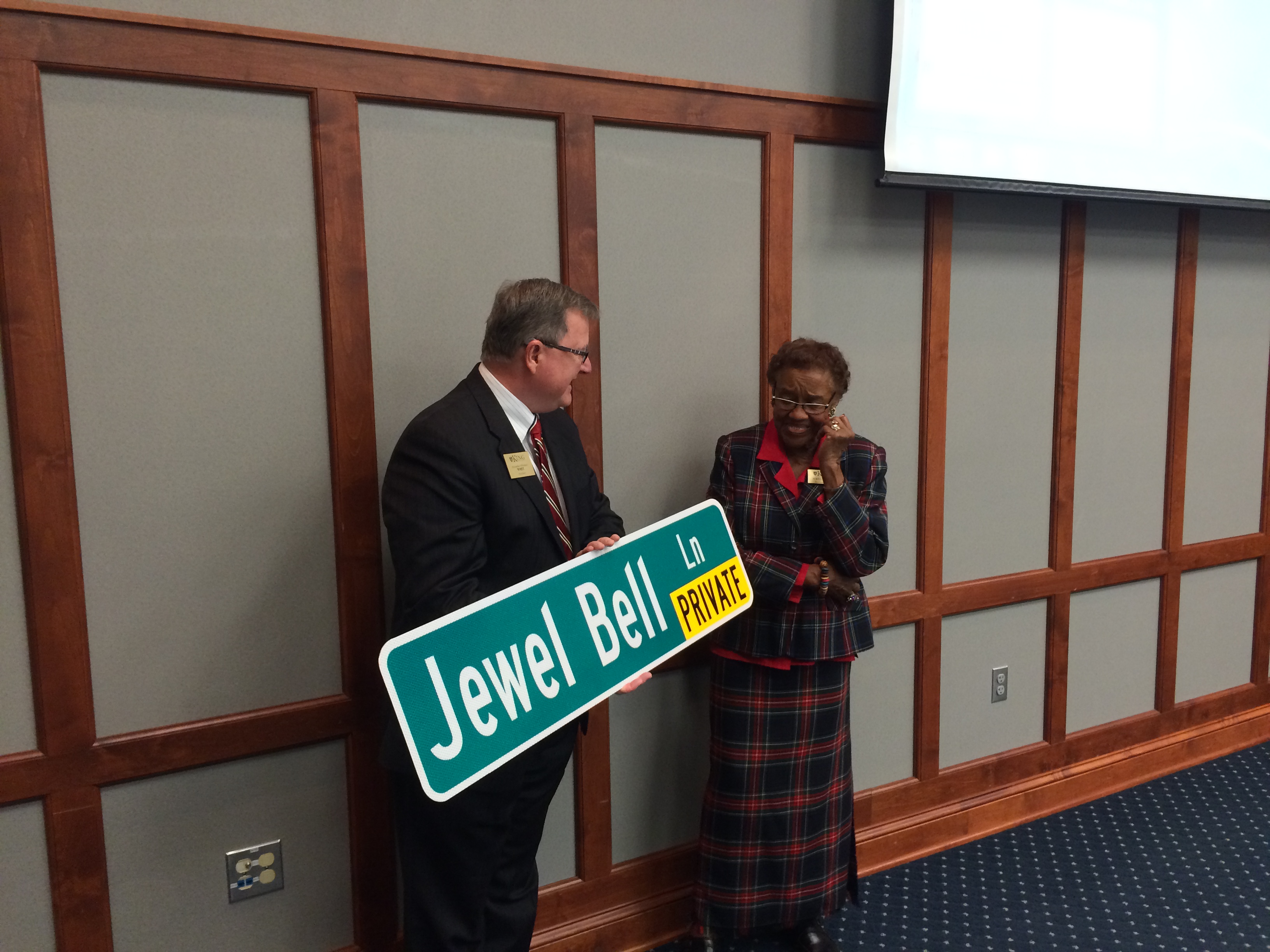 The King president, a white man wearing glasses, holds a street sign that says "Jewel Bell Lane" next to Bell, a Black woman with short hair wearing a plaid suit.